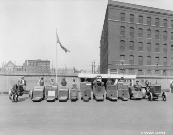 View towards a group of men posing outdoor on industrial tractors in the yard of what may be an International Harvester plant. There is a flag flying on a flagpole. The signs on the wall behind them read, in part: "Base-Ball, McCormick Safety Team." Buildings are in the background behind the wall.