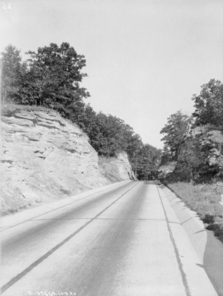 View down road. Rock has been blasted away to form the roadway. Trees are on top of the rock formations on both sides of the road.
