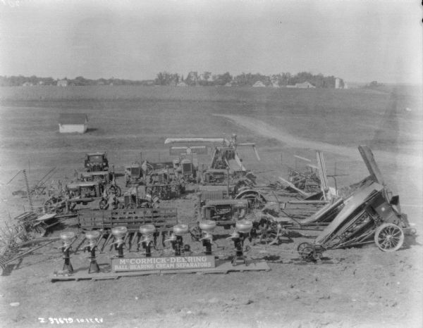 Slightly elevated view of farm implements on display outdoors in a field. A sign in front reads: "McCormick-Deering Ball-Bearing Cream Separators." There are also agricultural implements, a truck, and tractors on display. In the far background are farm buildings among trees.