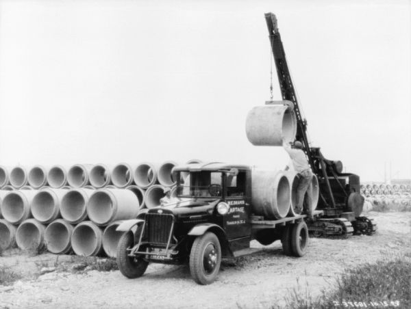 View towards a truck being loaded with large pipe from a crane. A man is helping to position the large pipe onto the back of the truck. The sign painted on the truck reads: "W.E. Draper Cartage"