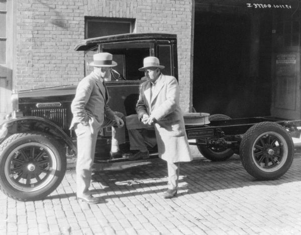 Two men are standing on the driver's side of a truck, each with one foot up on the running board. Behind them is a brick building with a sign that reads: "International Motor Trucks."