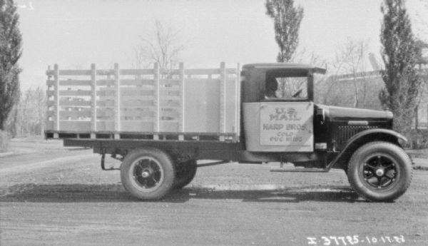 Right side view of a man sitting in the driver's seat of a truck with a stake body parked on a rural road. The sign on the passenger side door reads: "U.S. Mail, Harp Bros. Colo."