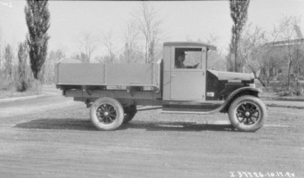Right side view of a man sitting in the driver's seat of a truck with a stake body parked on a rural road. There is a building behind trees on the right.
