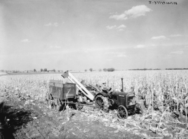 Elevated view towards a man driving a tractor pulling a corn picker in a field. A wagon is next to the corn picker.