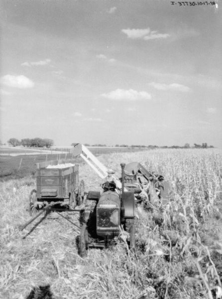 Elevated view from front of a man using a tractor to pull a corn picker in a cornfield. There is a wagon on the left next to the corn picker.