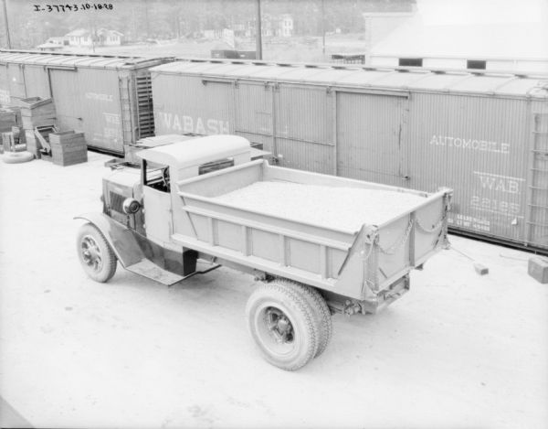 Elevated view of a dump truck on a railroad platform. Railroad cars are behind the truck, and buildings and trees are in the background.