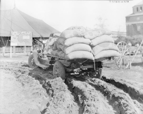 Rear view of the "Sahara" truck, with the back heavily loaded with sandbags, driving through deep sand at an exhibition. There is a map on a signboard in the background in front of a large tent on the left. There is a building in the background on the right.