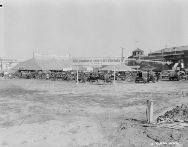 View across fairgrounds towards an International Harvester Company of America sign in front of a display area. Agricultural machinery and tractors are on display, and there are two tents. In the background on the right is a large building. The banner on top of the tent reads: "International Harvester Exhibit, McCormick-Deering Power Farm Equipment."