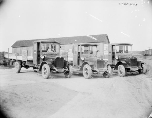 View towards three lumber delivery trucks parked in a row. The signs on the truck reads: "W.H. Chapman & Son." Railroad cars on railroad tracks on the right.