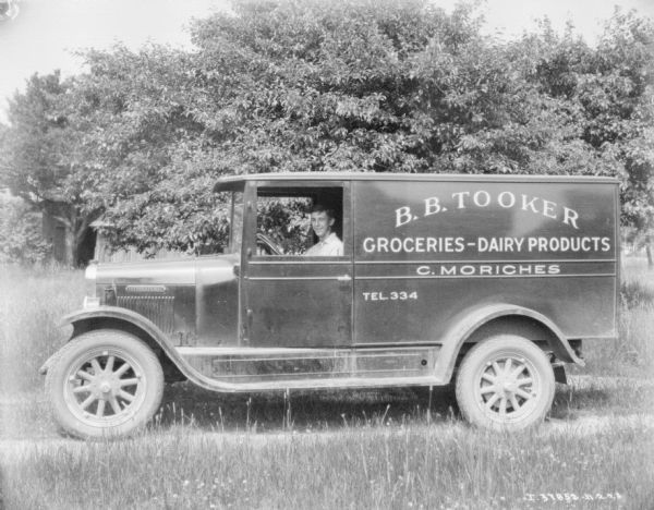 View towards a man sitting in the driver's seat of a delivery truck. The sign painted on the side of the truck reads: "B.B. Tooker, Groceries — Dairy Products, C. Morches."