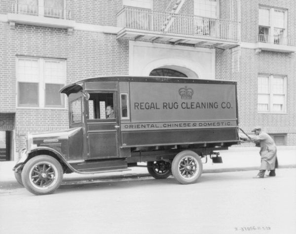 View across street towards a man sitting in the driver's seat of a delivery truck. A man is standing at the back of the truck, either removing or putting in a rug. The sign on the side of the truck reads: "Regal Rug Cleaning Co." Behind the truck is a large brick building.