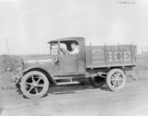 View across road towards a man sitting in the driver's seat of a delivery truck. The sign painted on the truck reads: "Nuzzi & Lorusso, Hygeia Ice."