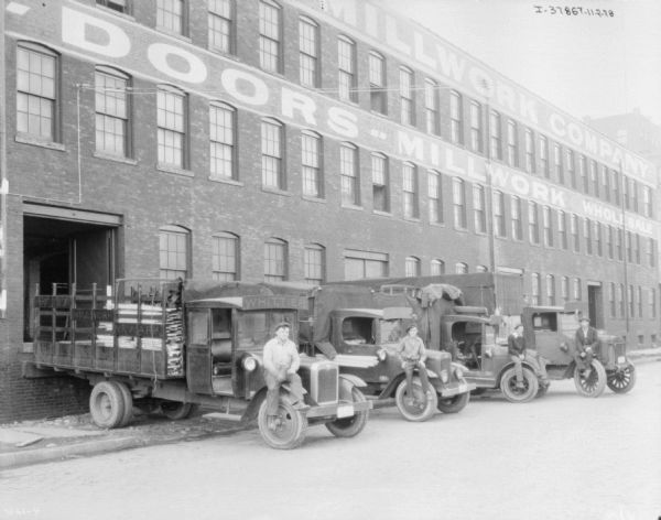 View towards four delivery trucks lined up in a row, all backed up against a brick building. The four men are posing sitting on the front right fender of each truck. The truck on the far left has signs that read: "Whittier," and Whittier Millwork Newark," and the truck is backed up to the loading dock of the building. The sign painted on the building reads, in part: "Millword Company, Doors — Millwork."