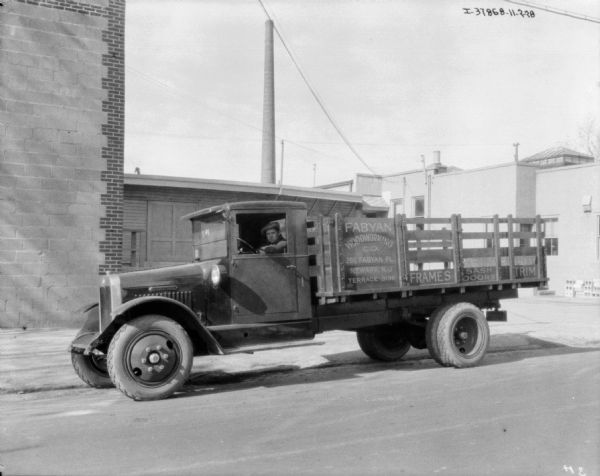 View across street towards a man sitting in the driver's seat of a delivery truck. The sign on the side of the truck reads: "Fabyan Woodworking Co., 266 Fabyan Pl., Newark, N.J." Behind the truck are industrial buildings.
