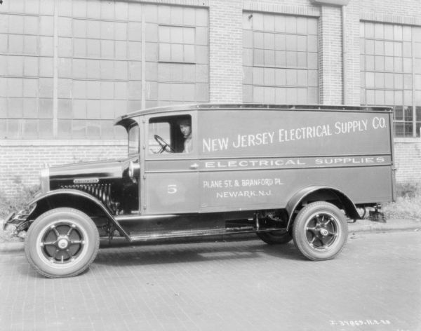 View across street towards a man sitting in the driver's seat of a delivery truck. The sign on the side of the truck reads: "New Jersey Electric, Electrical Supplies, Plant St. & Branford Pl., Newark, N.J." Behind the truck is a brick building with large windows.