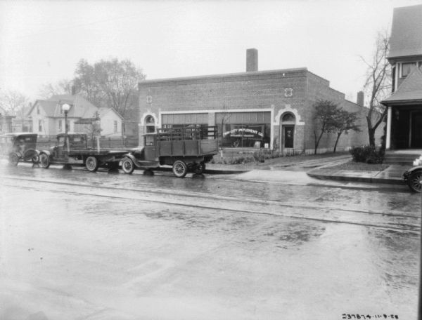 View across street towards a dealership. Trucks are parked along the curb in front of the building. The sign painted on the window reads: "Twin-City Implement Co., McCormick-Deering."