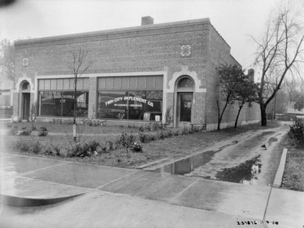 View from street towards the front of a dealership. The sign painted on the window reads: "Twin-City Implement Co., McCormick-Deering."