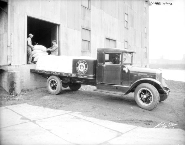A flour delivery truck is backed up to a loading dock. Two men are working with full flour sacks on a cart in the open doorway. The sign on the truck bed reads: "Royal Household, The Ogilvie Flour Mills Co. Limited."