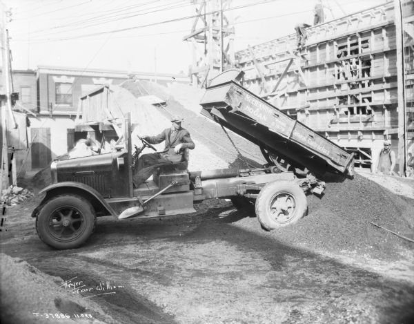 A man is dumping a load of coal from a coal delivery truck. A man is standing in the background on the right. The name painted on the side of the truck reads: "McCranor & Hogan, General Contractors."