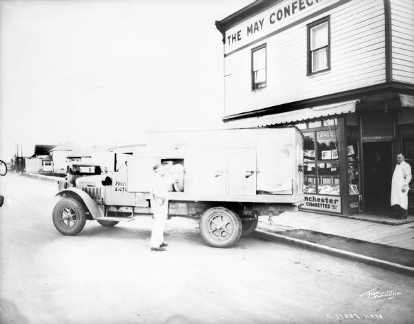 A man is making a delivery at small store. The sign on the building reads: "The May Confe[cut off]." A man wearing an apron is standing in the doorway of the storefront which has goods displayed in the front window.