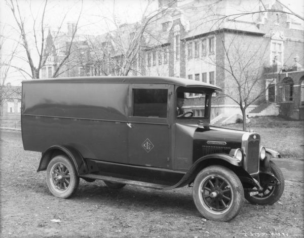 View towards passenger side of an enclosed truck. A man is sitting in the driver's seat. A Quebec license plate is on the front of the truck. In the background is a large brick building. A logo on the passenger door reads: "LC."
