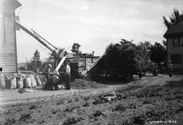 Three men are working with a McCormick-Deering thresher. A wagon piled with hay is on the right, and behind the wagon is a Farmall tractor.