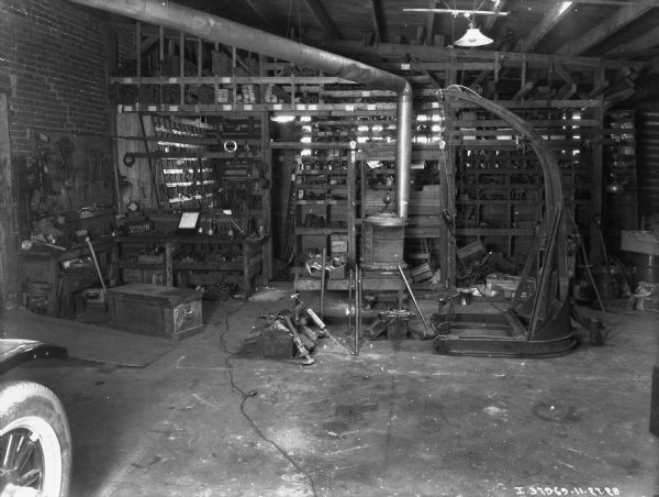 View of the interior of a repair shop. A wood burning stove is in the center, and tools are hanging on a board on the wall on the left. Parts are on shelves in the background. The wheel of a vehicle is in the left foreground.