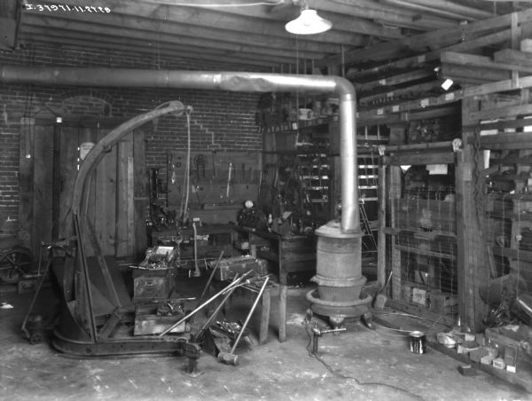 View of the interior of a repair shop. A wood burning stove is in the center, and tools are hanging on a board on the wall on the left. Parts are on shelves in the background.