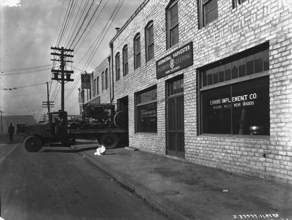 View down street towards a man sitting in the driver's seat of a truck backed up into the open garage door of the Evans Implement Co. There is a tractor on the bed of the truck. A sign hanging in the background reads: "Goodrich Silvertowns, A-1 Vulcanizing Works."
