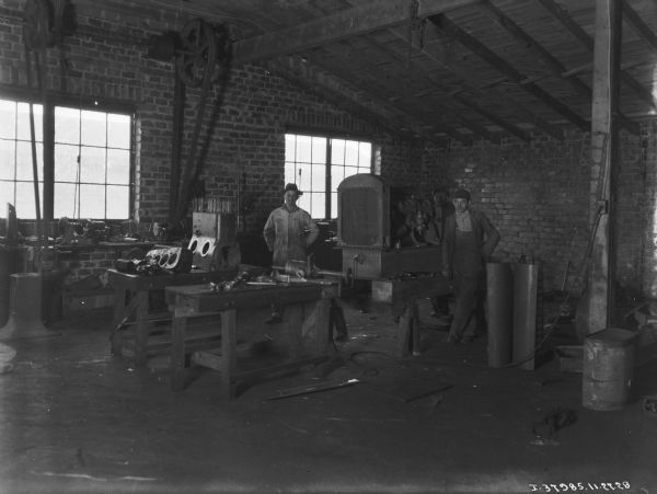 Three men are standing in a repair shop at a dealership. A tractor is in the center of the room.