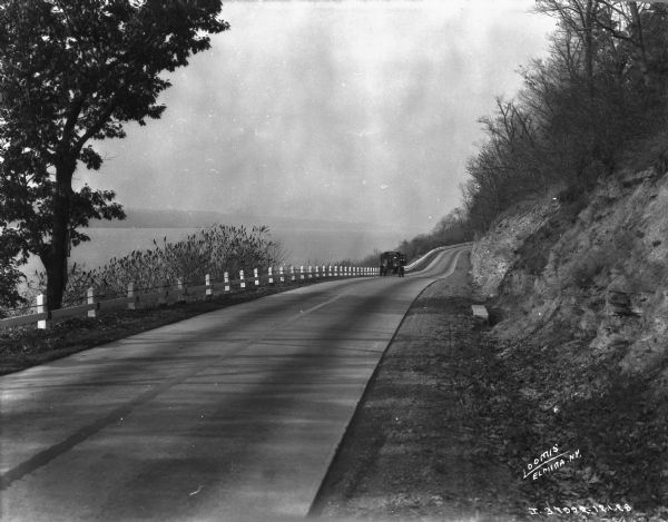View down highway towards a truck coming up the road. There is a long guardrail on the left, with a steep bank rising up pon the right. The highway is running along a large body of water, perhaps a river. The far shoreline is in the background on the left.