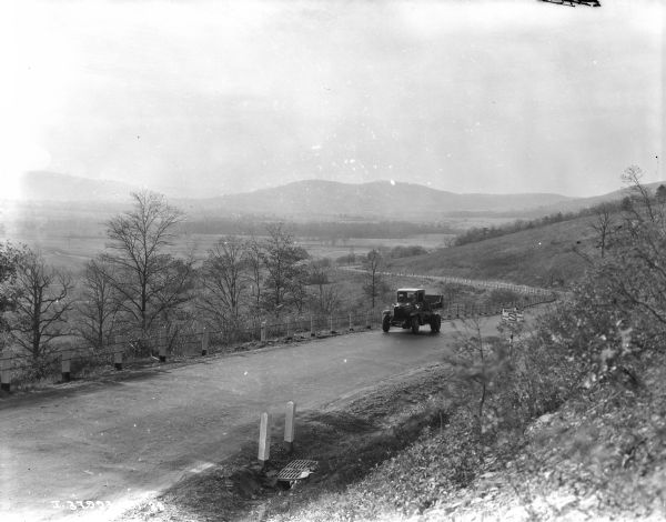 View down highway towards a truck coming up the hilly, windy road. There is a long guardrail on the left, with a steep bank rising up on the right. The highway is running along a valley, within hills in the background. A sign on the right side of the road reads: "Warning Sharp Curves."