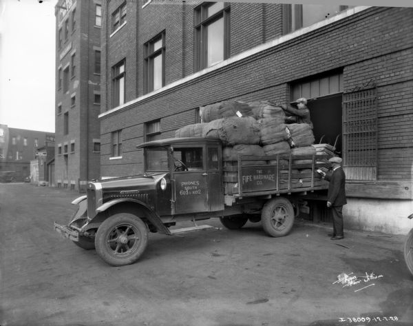 A truck is backed up to the loading dock of a large, brick building. The sign on the side of the truck reads: "The Fife Hardware Co." A man wearing a suit and hat is standing at the back left of the truck, which has a stake body and is piled with burlap sacks higher than the cab of the truck. Another man is standing on the loading dock, either loading or unloading the sacks.