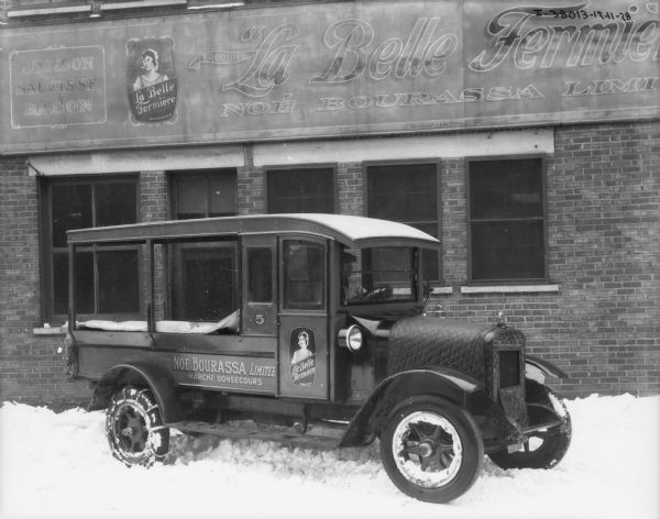 View towards a man sitting in the driver's seat of a truck parked in front of a storefront in Canada. On the front of the truck, and on top of the hood is a quilted, snap-on cover. The sign on the passenger side of the truck reads: "Noe Bourassa Limitée Marché Bonsecours." The same sign is on the brick building in the background. There is snow on the ground, and snow chains are on the rear tires of the truck.