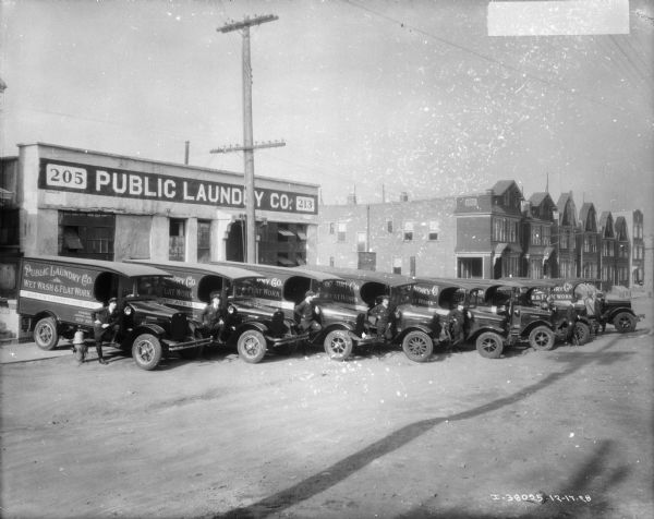 View down road towards a row of nine delivery trucks. Men in uniform are posing, some with their leg resting on the running board, of the passenger side of each truck. The building behind the group has a large sign that reads: "Public Laundry Co. Inc." A row of apartment houses is in the background.