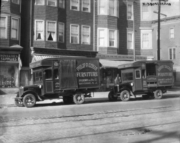 View across street towards two delivery trucks, one for Philip P. Cohan Furniture, and one for Harry Sacks Furniture, both of Bayonne, N.J. parked along the curb. There are storefronts with apartments above behind the trucks. A storefront with a show window on the left is advertising "Novelties Supplies."