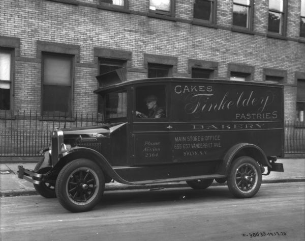View across street towards a man sitting in the driver's seat of a truck. The sign on the side of the enclosed truck reads: "Finkeldey Bakery Cakes Pastries, B'klyn, N.Y." A large brick building is behind an iron fence in the background.