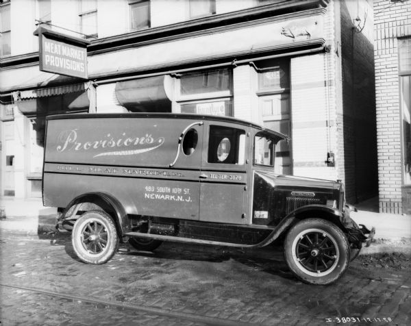 View across cobblestone street towards an enclosed delivery truck. The sign on the side of the truck reads: "Provision's Hill Meat Market, Inc." The building behind the truck has a sign that reads: "Meat Market Provisions," and a decoration above the window at the corner of the building on the right side is of a sow.
