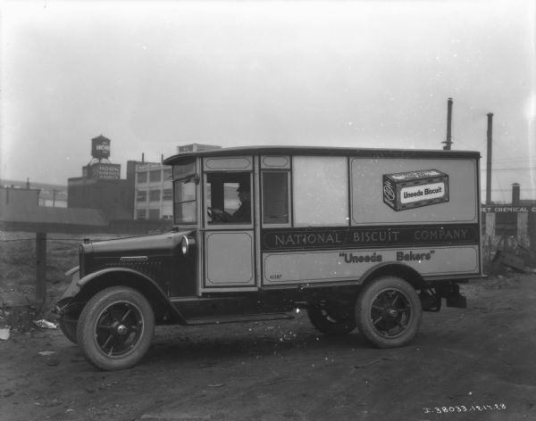 View towards a man sitting in the driver's seat of an enclosed truck parked on an unpaved road. The sign painted on the side of the truck reads: "National Biscuit Company, 'Uneeda Bakers.'" Industrial buildings are in the far background.