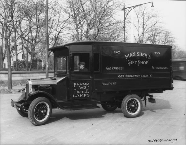 View towards a man sitting in the driver's seat of an enclosed truck parked on an unpaved road. The sign painted on the side of the truck reads: "Go To Max Sher's 'Gift Shop.,' Gas Ranges, Refrigerators, Floor and Table Lamps."