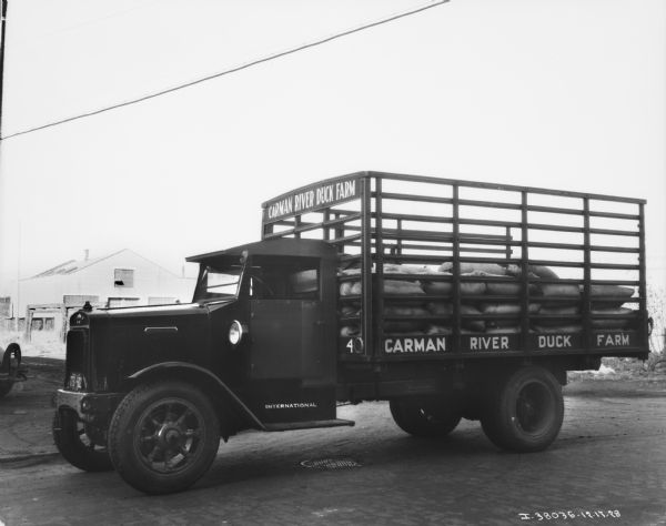 View towards the driver's side of a delivery truck with a stake body. The sign on the truck reads: "Carman River Duck Farm." There are filled burlap sacks piled in the back of the truck. Industrial buildings are in the background.