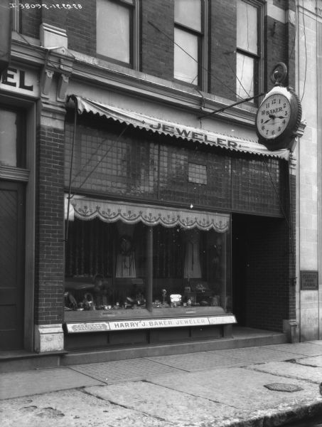View from street towards the front of a jewelers shop. There is a large clock hanging above the sidewalk. The sign below the show window reads: "Harry J. Baker, Jeweler." The building next door on the right has a plaque that reads: "Peoples State Bank."