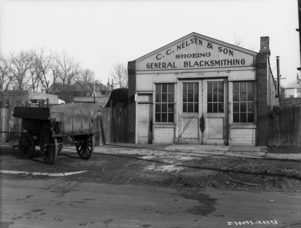 View across road towards a blacksmith shop. The sign on the front reads: "C.C. Nelsen & Son Shoeing General Blacksmithing." A wagon is parked on the left.