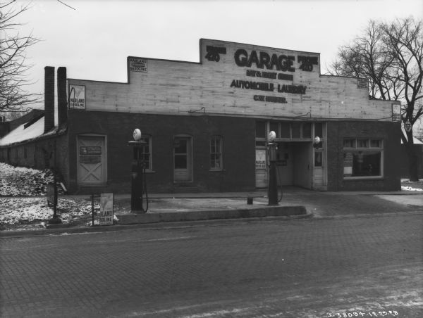 View across cobblestone street towards a building with a sign that reads: "Garage, Day & Night Service, Automobile Laundry, C.W. Huber." There are gas pumps at the curb. On the building are signs for Marland Gasoline, Vesta Batteries, and Pennnzoil.