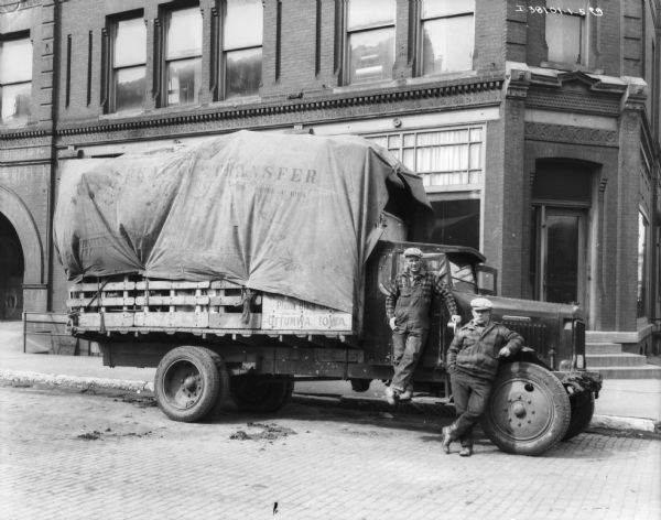 Two men are standing with a moving van parked along a curb. The back of the truck is covered with a tarp. A large brick building is behind them.