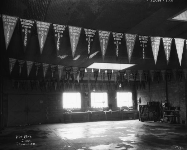 The IH banners hanging from the ceiling read: "McCormick-Deering Farm Equipment." There are skylights in the ceiling, and windows on the back wall are over work tables. Tool racks are along the brick wall on the right. A set of double doors are on the left.