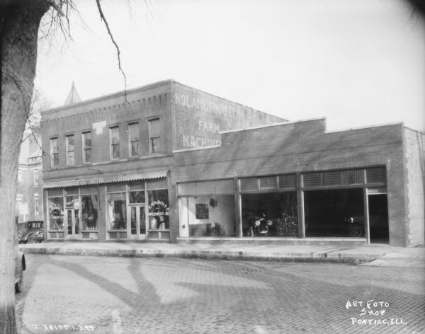 View across cobblestone street towards a dealership. There are show windows along the sidewalk, and the sign painted on one of the windows reads: "E.M. DeBolt Implement Co."