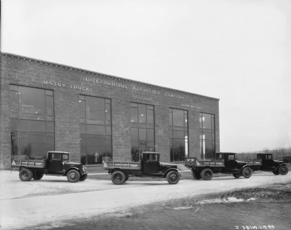 Four trucks are parked outside a large dealership. The signs painted on the trucks reads: "Allen County Highway Department." The two-story brick building in the background has a sign that reads: "International Harvester Company of America, Motor Trucks, Tractors, Farm Machines." Men are standing inside looking out of the floor-to-ceiling windows of the dealership.