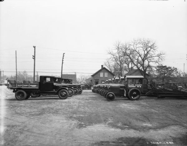 View towards two rows of trucks parked in a lot facing each other. The signs painted on the trucks reads: "Allen County Highway Department." Men are sitting in the trucks parked on the left. Buildings are across the street in the background.