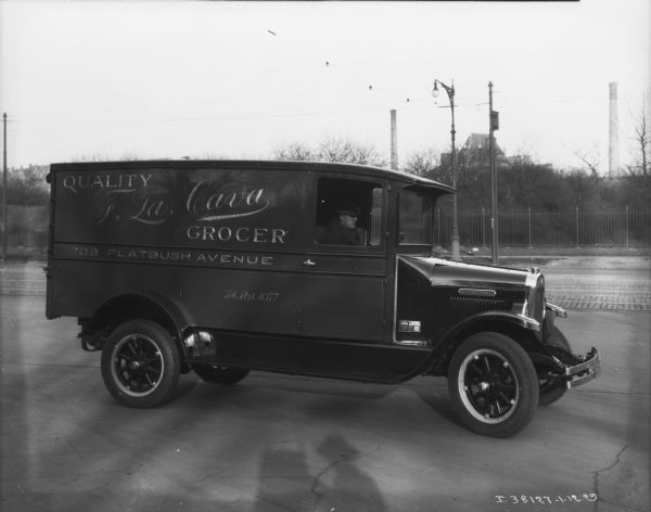 View towards the passenger side of a grocery delivery truck. A man is sitting in the cab. The sign on the side of the enclosed truck reads: Quality F. Lsa. Cava Graocer."